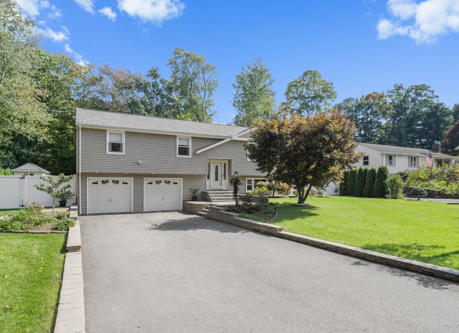 Gray raised ranch home with an attached two-car garage, a white fence, and a landscaped front yard with a tree in Farmington CT