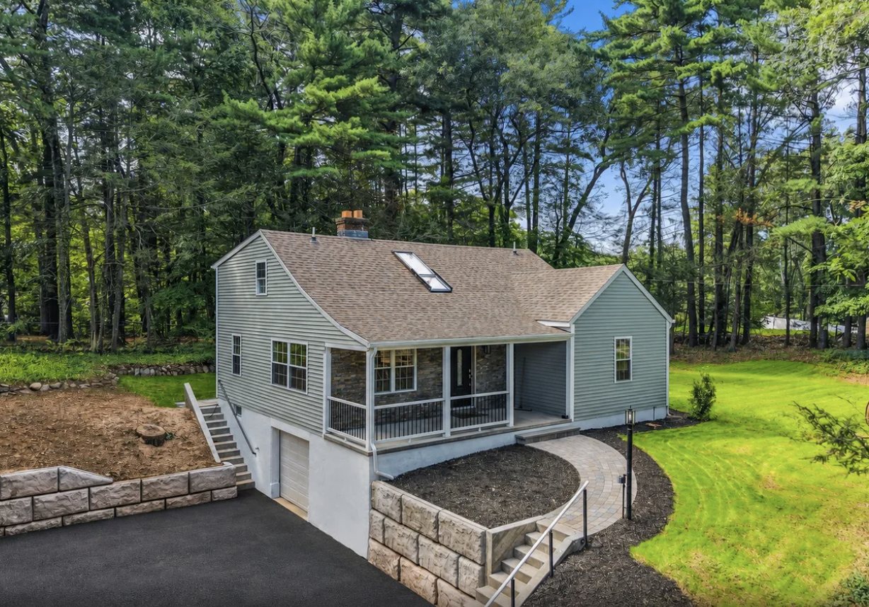 Renovated gray cape-style home with a covered stone-faced porch, a skylight, a lower-level garage, a paver walkway, and stone block retaining walls on a wooded lot