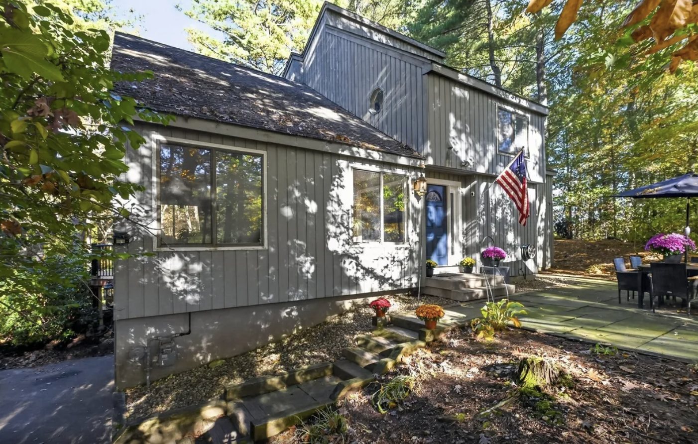 Gray contemporary home with vertical siding, a blue front door, an American flag, a stone patio with outdoor seating, and a wooded lot in autumn