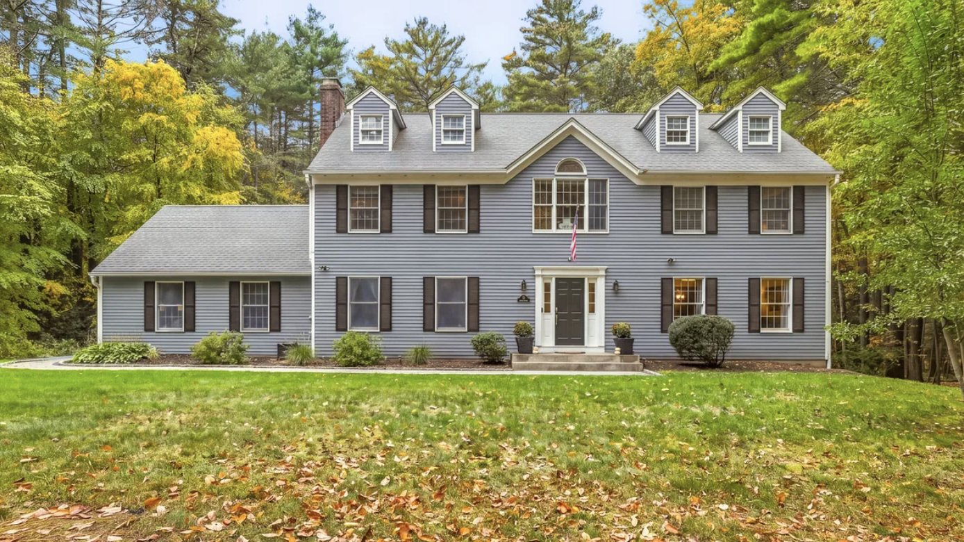 Two-story gray colonial home with black shutters, three dormers, an attached garage, and a wooded lot in autumn in Simsbury CT