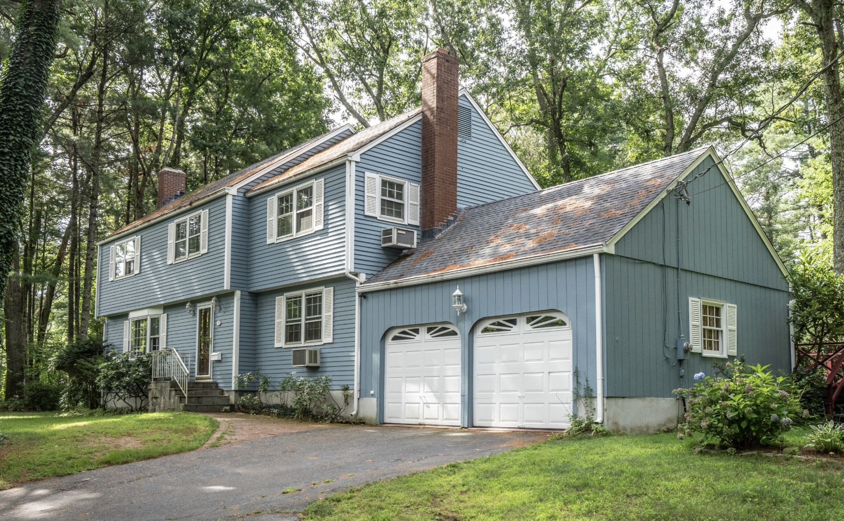 Two-story blue colonial style home with white shutters, a brick chimney, an attached two-car garage, and a wooded lot in Simsbury CT