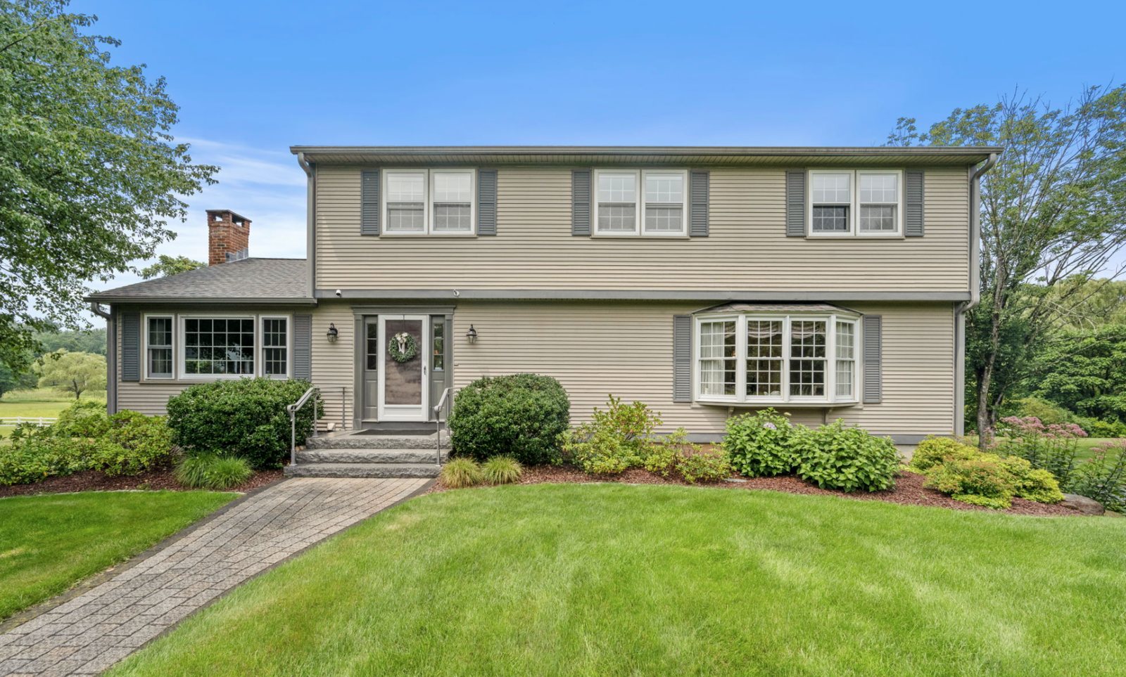 Two-story beige colonial home with gray shutters, a bay window, a brick chimney, granite front steps, and a landscaped lawn in Simsbury CT