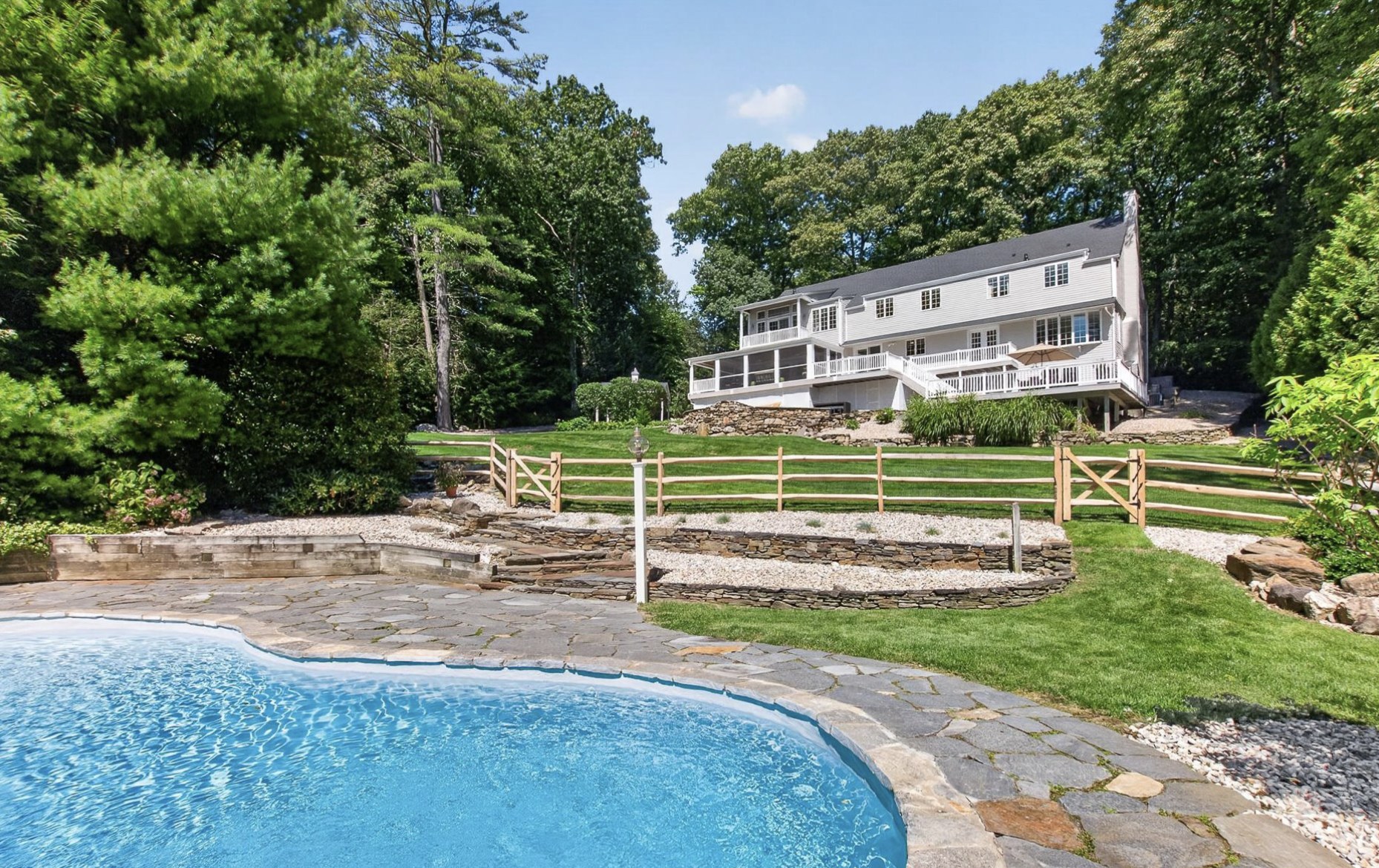 Rear view of a large home with decks and a screened porch above a stone terraced yard and a freeform swimming pool in Simsbury CT